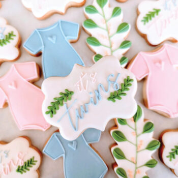 Cookies decorated like onesies in pink and blue with a cookie on top saying It's Twins