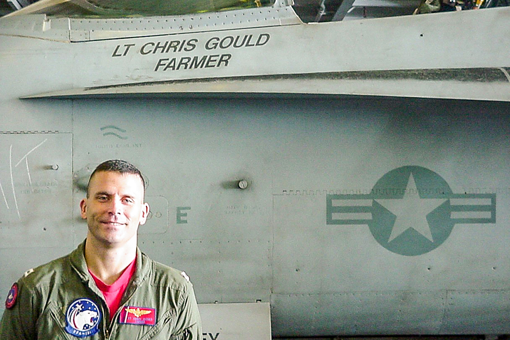 Chris Gould in his pilot uniform with the plane behind him that has his name and Farmer call sign