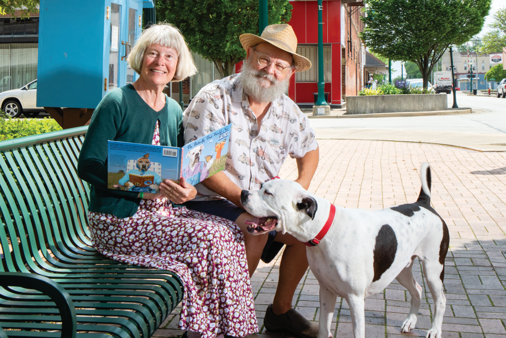 Kirby and Cindy sitting on a bench holding one of their books with their dog, Joe, an inspiration for one of their "dog people" Dogtown Artworks